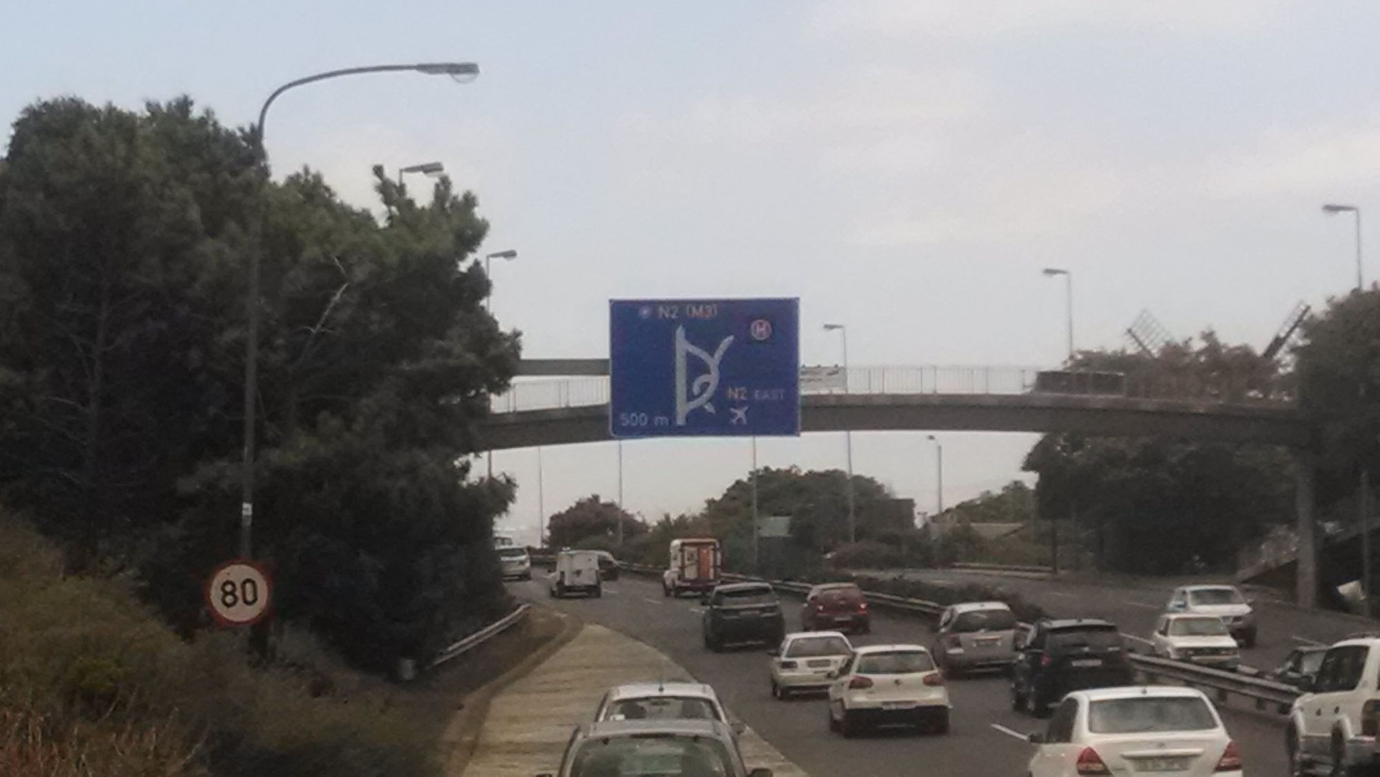 Cars are driving on a multi-lane highway under an overhead pedestrian bridge. There is a blue road sign above.